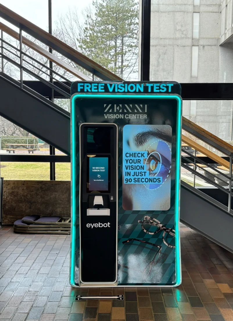 Free Vision Test kiosk installed in a building atrium, its touchscreen reading 90 second vision test, tap to get started.